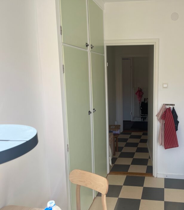 Kök med grönt skåp och trästol, öppning till lång korridor med rutigt golv och dörrposter utan dörrar; redskap hänger på väggen.
