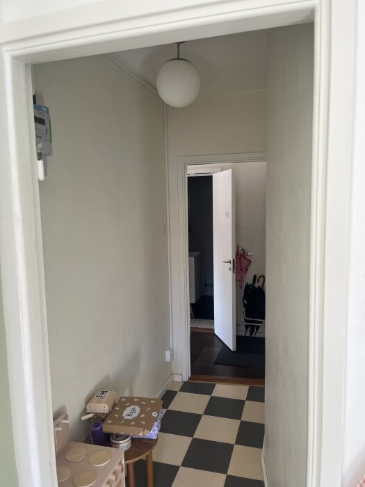 Lång, smal korridor med schackrutigt golv, ett bord längs ena väggen och en öppen dörr vid korridorens slut, lampa i taket.