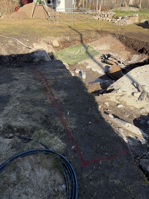 Grävt område med jord och stenar, markerad linje och liggande blå kabel, omgiven av gräs. Grön presenning och grushög i bakgrunden.