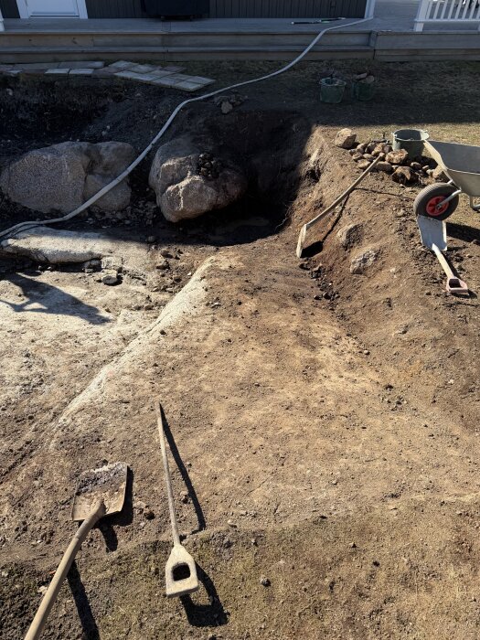 Grävt område med spade, spett och skottkärra i förgrunden, stora stenar och en träterrass i bakgrunden.