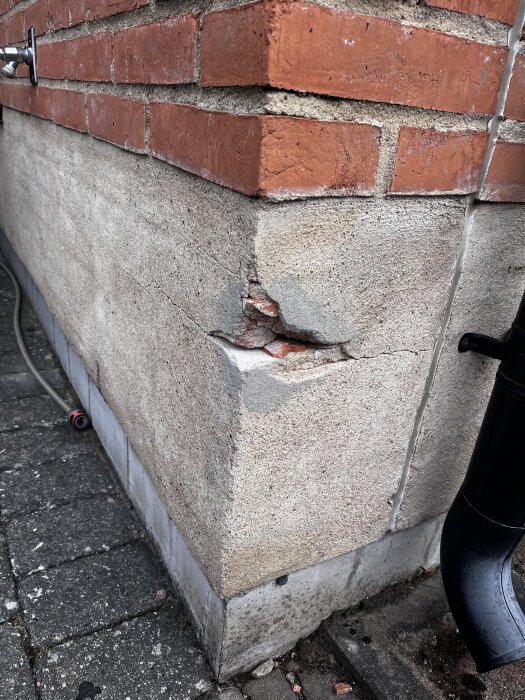 Skadad hörna på en röd tegelvägg med synliga sprickor och en tidigare lagning i putsen, omgiven av en betongplatta.