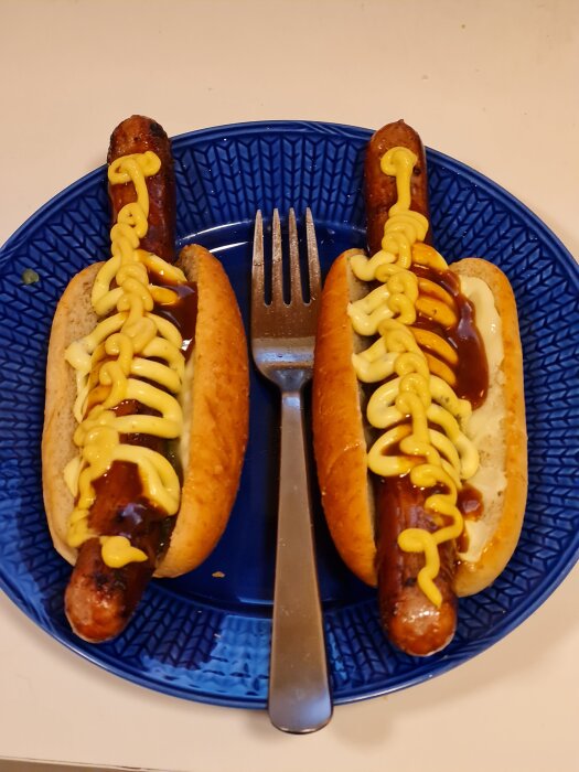 Två korvar i bröd med senap och sås på en blå tallrik, gaffel mellan korvarna.