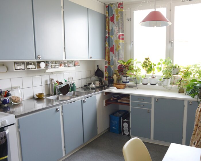 Kök med blågrå funkisstilsluckor, rostfri diskbänk, vita kakelplattor och växter på fönsterbrädan. 1950-60-talsinredning.