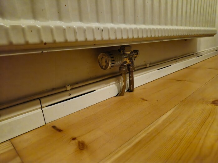 Radiator med anslutande rör och plastsockel nära golv, belägen vid en yttervägg, med ventilerad list synlig på trägolv.