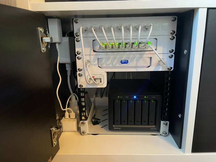 Öppet skåp med nätverksutrustning, inklusive en NAS och switch, inkopplat med kablar och LED-lampor som lyser.