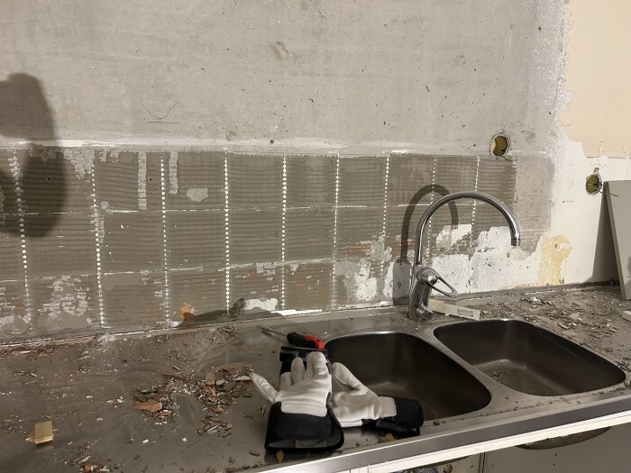 Köksområde under renovering, gamla plattor borttagna, vägg med kvarvarande kakelfix, verktyg och handskar på diskbänk.