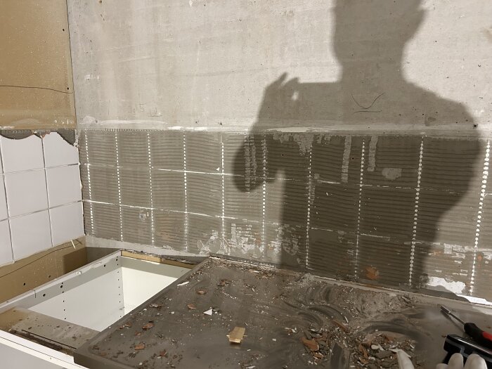 Kök under renovering med borttagna kakelplattor. Vägg täckt av kvarvarande kakelfix. Synliga verktyg och skugga av person på väggen.