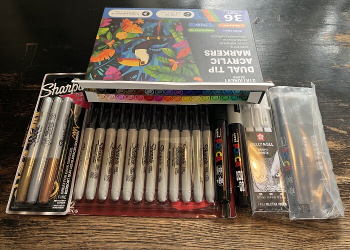 Olika sorters tuschpennor och markers på ett träbord, inklusive Sharpie och Uni Posca, samt akrylfärgspennor i en färgglad förpackning.