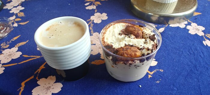 En kopp macchiato bredvid en efterrätt på ett bord med blommigt blått duk.