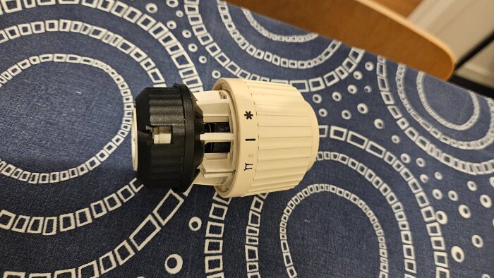 Gammal Danfoss radiatorventil på ett bord med mönstrad duk, vit och svart i färg, med temperaturinställningar synliga.