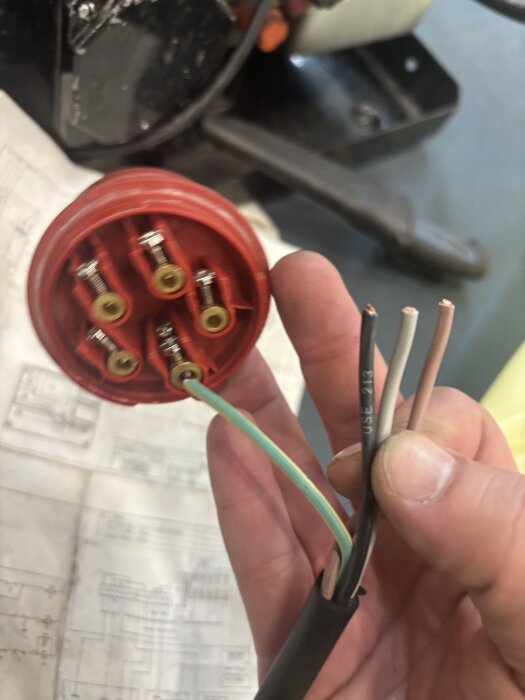 Hand som håller en CEE-kontakt med oskalad fyrledad kabel, i bakgrunden syns ett kopplingsschema.