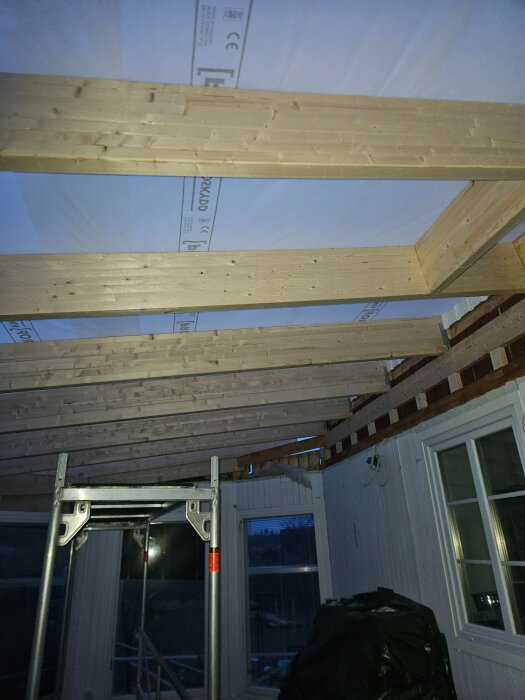 Trätakstolar i en pågående renovering av ett rum med vitmålade väggar och fönster. En byggställning syns i förgrunden.