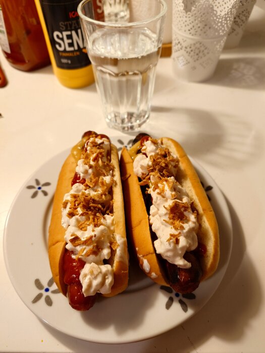 Två hot dogs med ketchup, senap, rostad lök och dressing på en tallrik, med ett glas vatten i bakgrunden.