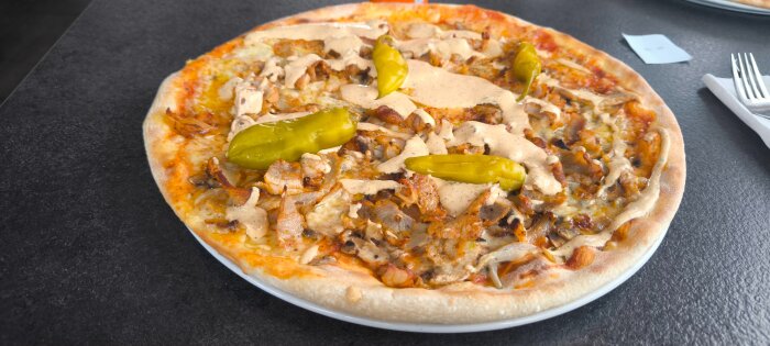 Pizza toppad med kebabkött, grön paprika och en krämig sås på en tallrik mot en mörk bordsyta.
