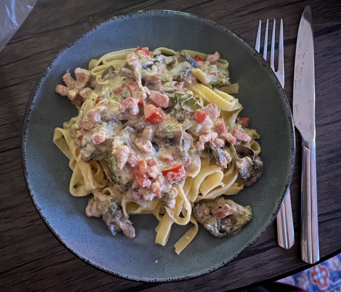 Tallrik med färsk pasta och pastasås gjord på bacon, broccoli, lök, paprika, champinjoner, svartpeppar och parmesan på ett bord med bestick.