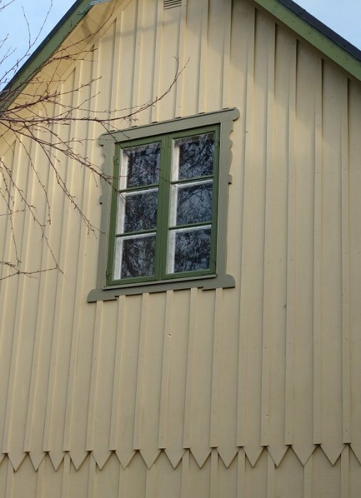 Gult trähus med grönt målat fönster, bilden visar en del av fasaden med liggande träpanel och detaljer kring fönsterkarm.
