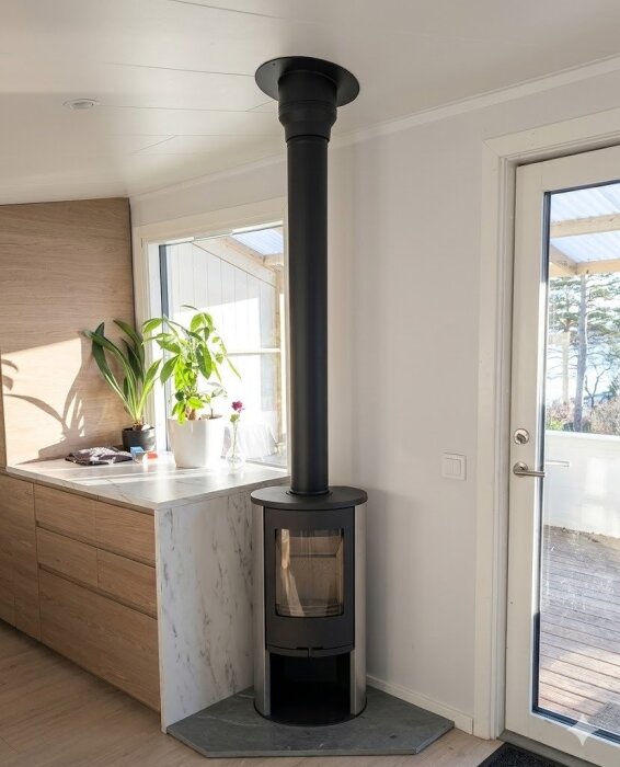 Svart kamin med rör som sträcker sig till taket, placerad bredvid köksbänk med några krukväxter, i ett rum med vita väggar och trägolv.