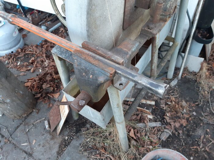Verkstad med skruvstäd som håller metallstång framför en slipsten, omgiven av verktyg, gammalt trä och höstlöv.