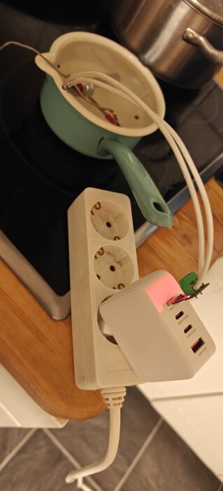 USB-C laddare kopplad till konsumentelektronik, en blå kastrull används ovanpå en induktionshäll, kabel draget in i kastrullen.