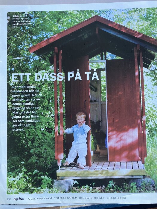 Ett barn står leende framför en röd traditionell utedass med dörrarna öppna, omgivet av grönska.