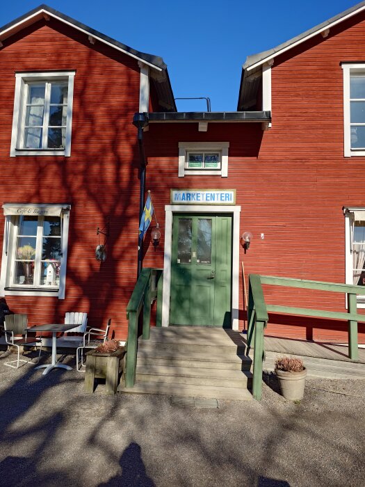 Röd byggnad med två våningar, grön dörr och skylt med texten "Marketenteriet". Uteservering med bord och stolar, svensk flagga vid entrén.