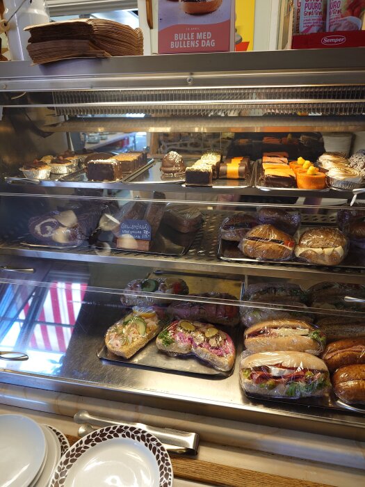 Förpackade smörgåsar och bakelser i en glasmonter på ett café, med en skylt för "Bulle med Bullens dag" synlig ovanför.