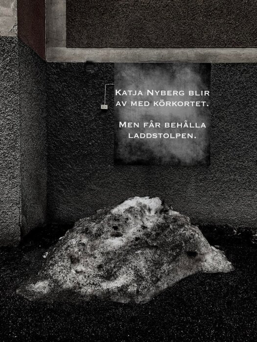 En skylt med text om Katja Nyberg, som förlorar körkortet, ovanför en smutsig snöhög vid en betongvägg.