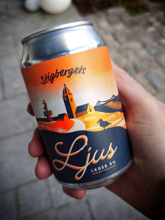 En hand håller i en ölburk med etiketten "Stigbergets Ljus Lager 5%", beige och orange design, utomhus på stengolv.