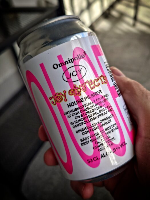 Bryggd burk öl Omnipollo Joy Objects House Pilsner hålls i handen utomhus, etikettens text och färg är synliga.