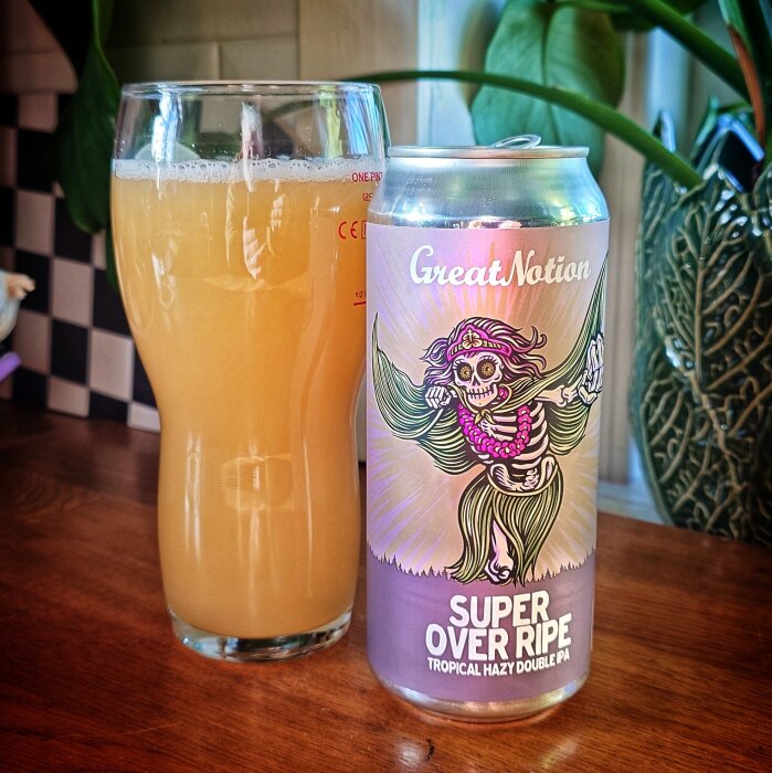 Burk med "Super Over Ripe" Tropical Hazy Double IPA och ett glas med gyllene öl på ett bord, på en bakgrund av plantor.
