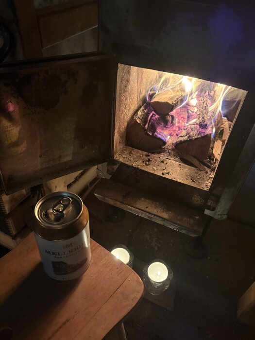 Öppen spis med eld och en burk Melleruds Pilsner står på en träbänk bredvid två tända ljus i en bastu.