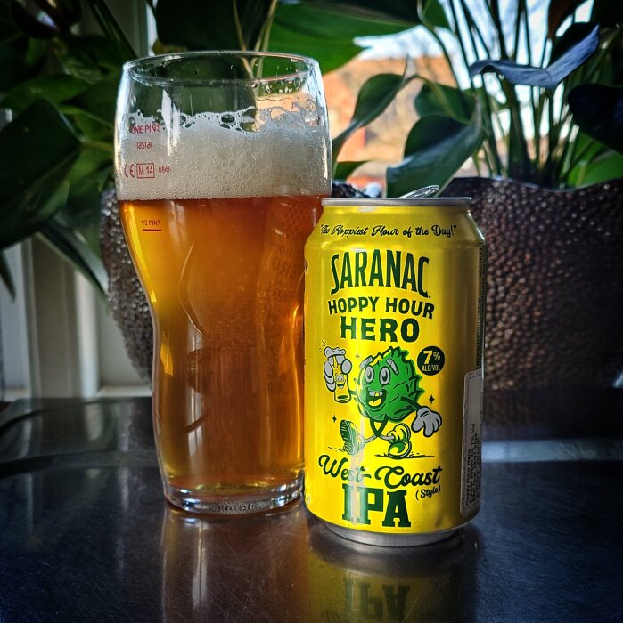 En glas öl bredvid en burk Saranac Hoppy Hour Hero West Coast IPA, omgiven av gröna växter.