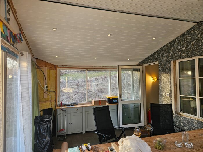 En verandao med isolering synlig på ena väggen, grå skåp, bord med föremål och utsikt mot en trädgård. Lampor i tak och på vägg.