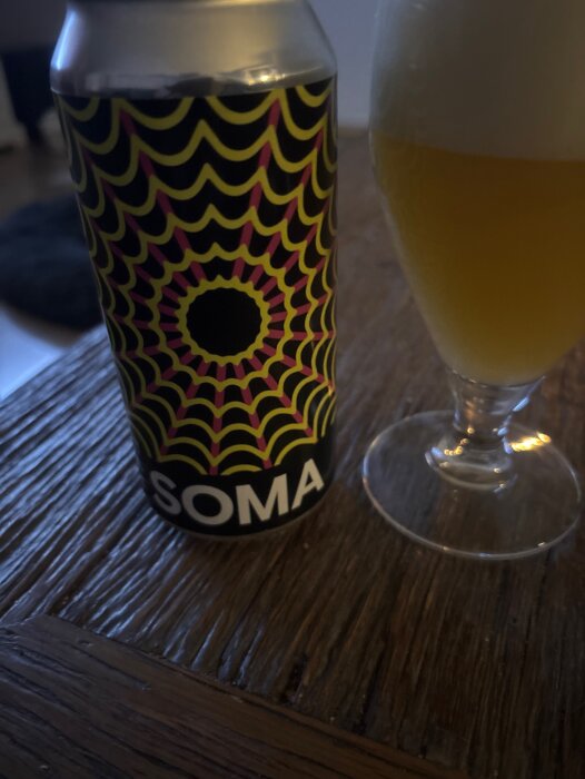En burk av Soma Nebula Dipa med ett färgglatt mönster, bredvid ett glas med gyllene öl på ett träbord.