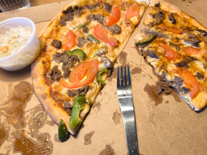Hämtpizza med tomat och paprika på kartong, bredvid en plastburk med kålsallad och en gaffel.