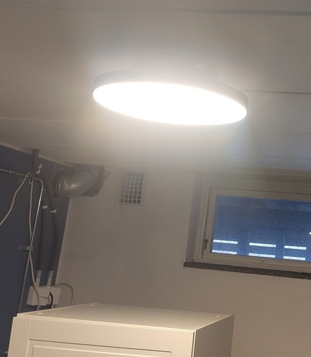 Taklampa lyser i ett rum med fönster, ventilationsledning och kablar på väggbredden.