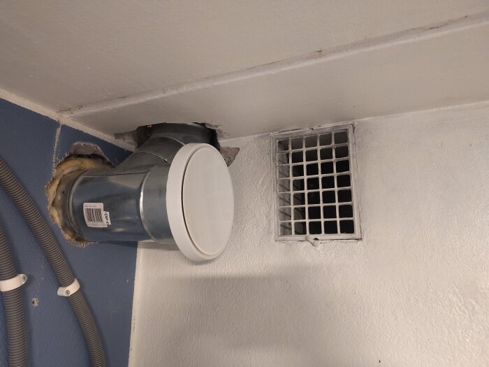 Ventilationstrumma i metall installerad i ett hörn, bredvid ett fyrkantigt luftgaller på en vit vägg, med grå plaströr vid sidan.
