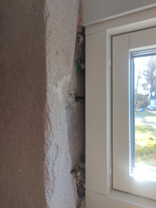 Vägg under renovering nära ett fönster med synlig öppning mellan murbruk och fönsterkarm. Soligt ljus utanför.
