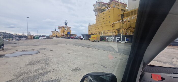 Hamnområde med gula fartyg och parkerad DHL-bil sedd genom bilruta.