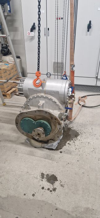 Industrikomponent på verkstadsgolv med kedjor och oljefläckar runtom.