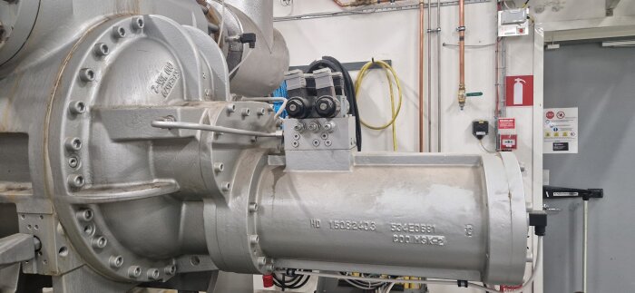 Industriell maskin i en verkstadsmiljö med rör och kablage synligt i bakgrunden.