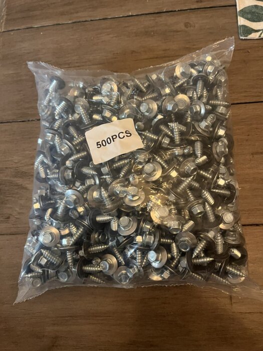 En påse med 500 stycken rostfria självgängande skruvar med gummitätning liggande på ett träbord.