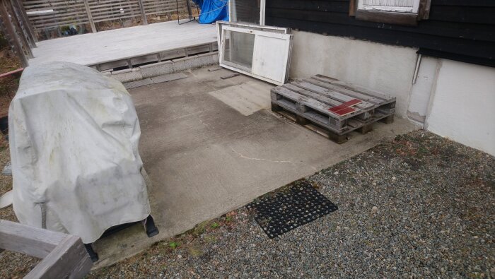 Betongplatta vid hus med två träpallar, ett fönster lutat mot väggen och en täckt struktur. Trall och grus synliga i bakgrunden.