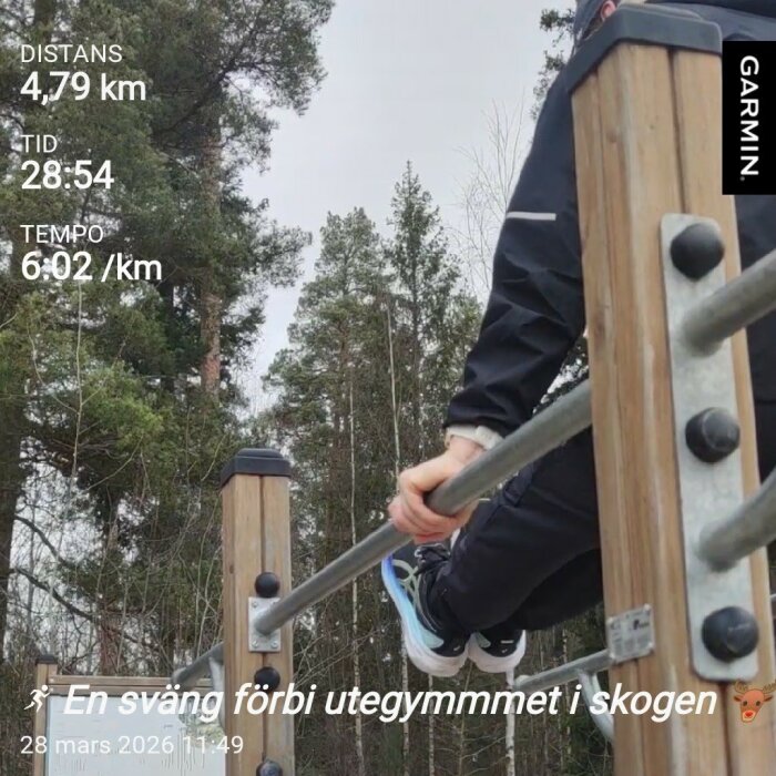 Person tränar på ett utegym i skogen med tröja och träningsskor, omgiven av höga träd. Text på bild visar distans, tid och tempo från ett löppass.