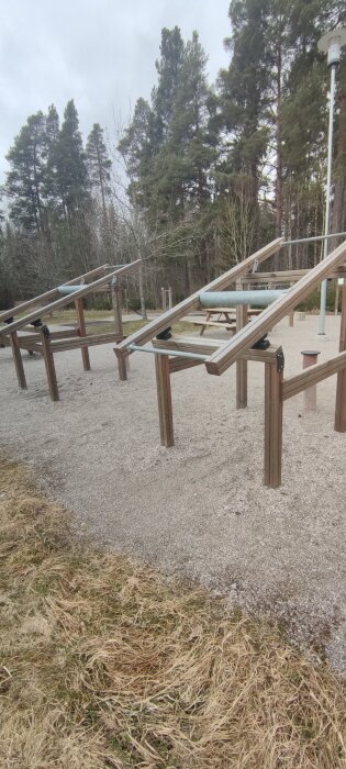 Utomhusgym med träningsställning av trä och metallrör på grusmark, omgiven av skog och bänkbord i bakgrunden.