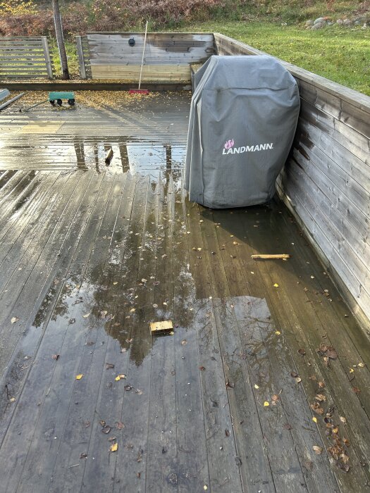 Träaltan med löv och vattenpölar, täckt grill i hörnet, kratta och leksak på golvet.