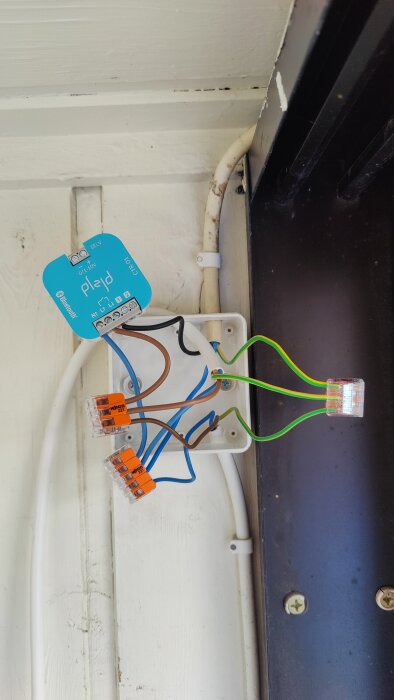 Elektrisk kopplingsdosa med flera färgkodade kablar och en blå enhet märkt med Bluetooth-teknik, monterad på en trävägg.