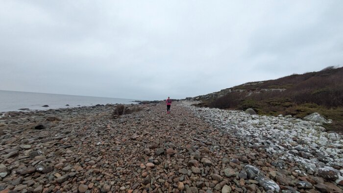 Person jogging längs en stenig kustlinje med havet till vänster, under en molnig himmel.