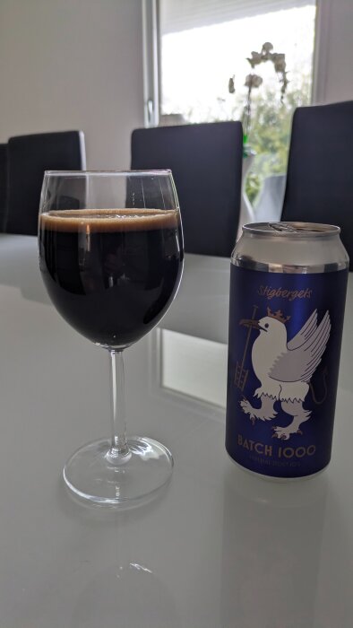 Svart öl i vinglas bredvid burk med blå etikett med vit fågel och texten "Batch 1000" på bord i matsal.