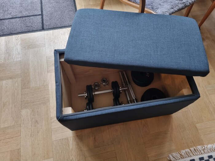 Blå sittpuff med avtagbart lock visar förvaring av hantlar och vikter på ett trägolv.